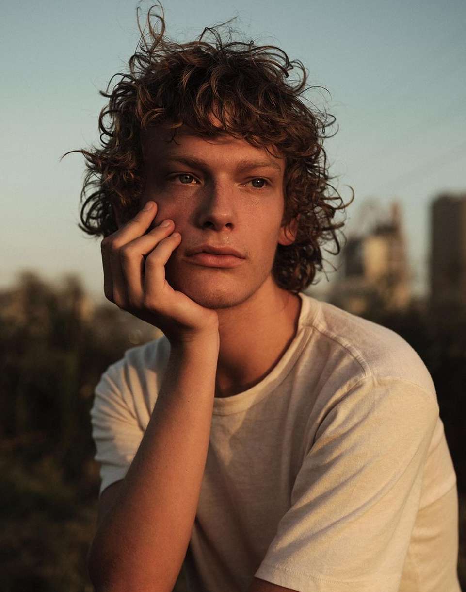 Portrait of a curly-haired young adult with hand on chin against an outdoor city background, bathed in golden hour sunlight.