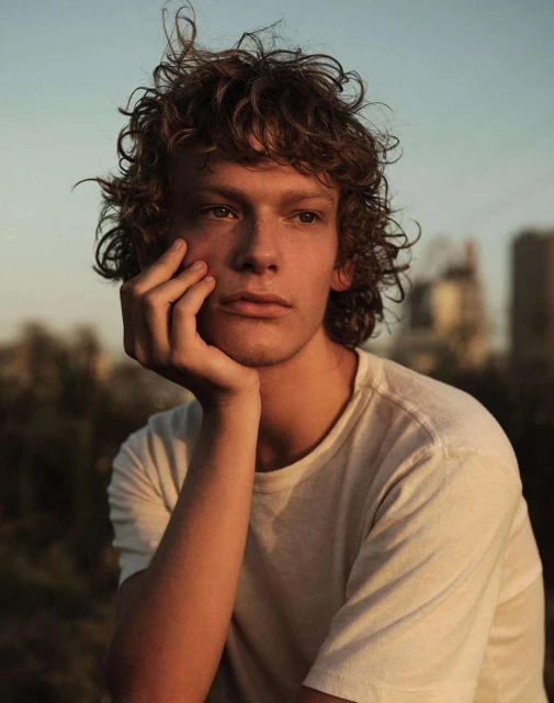 Portrait of a curly-haired young adult with hand on chin against an outdoor city background, bathed in golden hour sunlight.