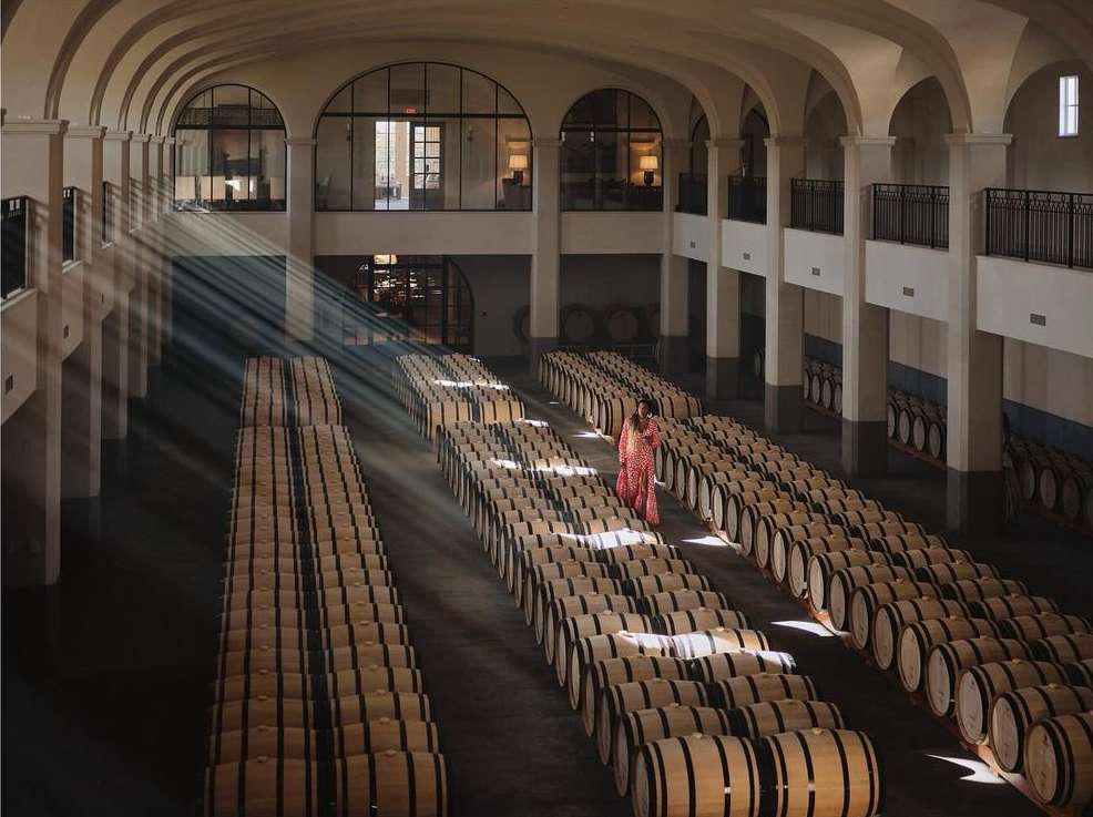 Large wine cellar featuring rows of barrels and a person in a red dress walking. Sunlight streams through the arched windows.
