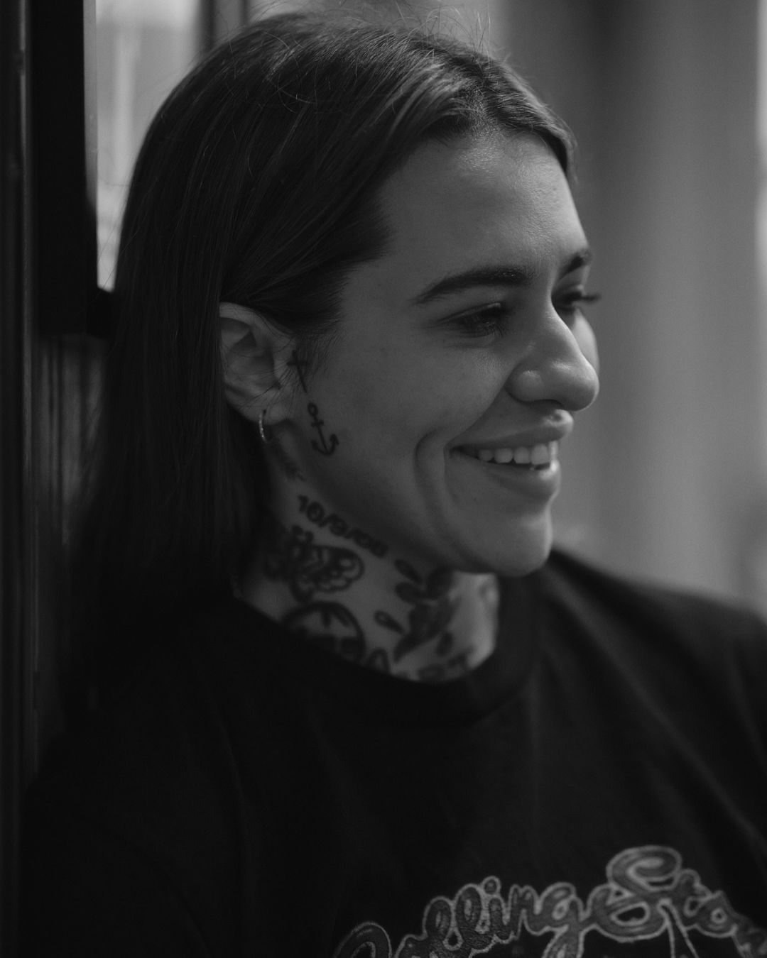 A black and white portrait of an adult woman with tattoos on her neck and face, smiling and wearing a dark crewneck shirt indoors.