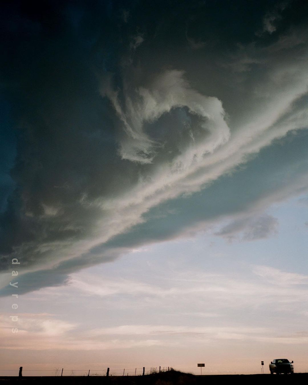 Dramatic storm clouds fill the frame above a rural highway with a vehicle driving into the distance at sunset.