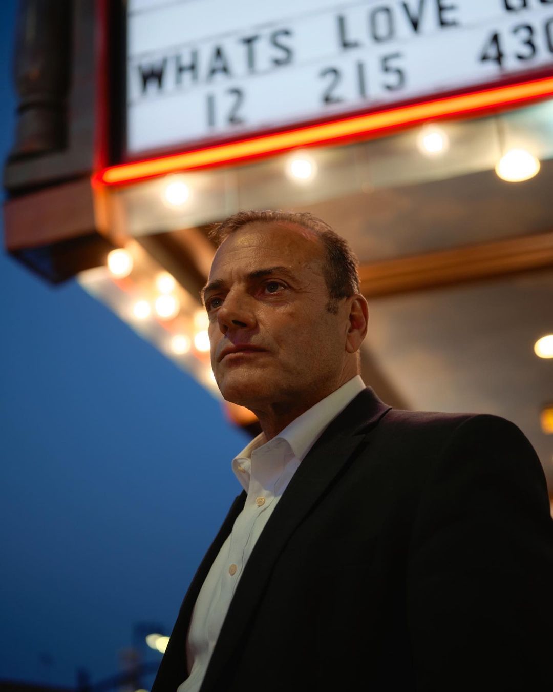 Portrait of a man in a suit standing in front of a theater showing movie listings at night, captured in a low-angle shot.