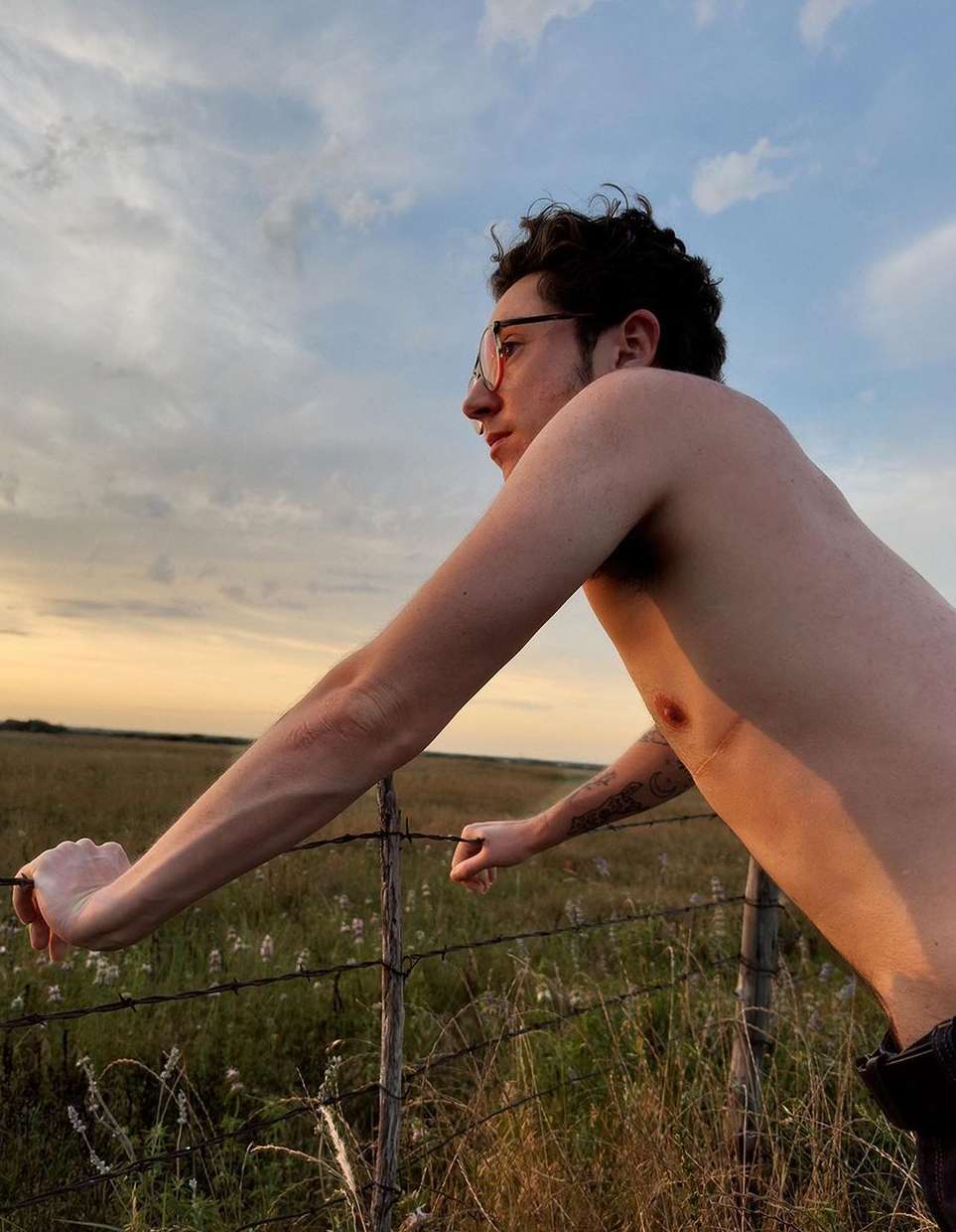Man leaning on fence in field at sunset with bare chest and glasses, looking away from camera, with a warm color palette.