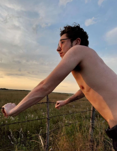 Man leaning on fence in field at sunset with bare chest and glasses, looking away from camera, with a warm color palette.