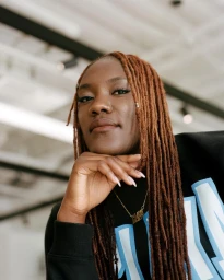 Portrait of a black woman with long locs resting her chin on her hand, wearing a black sweatshirt with light blue lettering.
