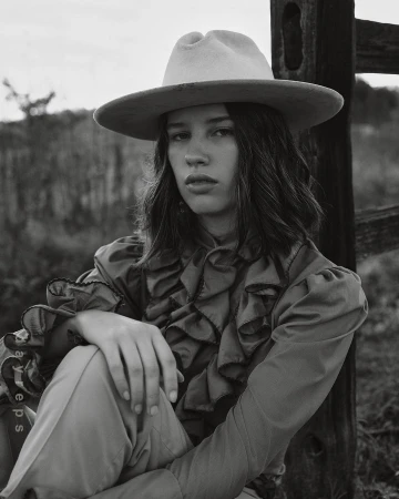 Black and white portrait of a woman wearing a wide-brimmed fedora hat and ruffled blouse outdoors near a rustic wooden fence.
