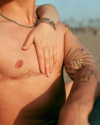Close-up of a shirtless person with arm tattoos and chain necklaces, with another person's hand resting gently on their chest