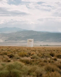 A white water tank featuring text stands in a dry, sparse desert landscape under a cloudy sky with distant mountains.