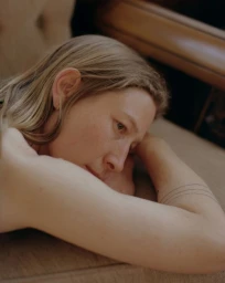 Close-up portrait of a woman with blonde hair resting her head on her arms, featuring fine line tattoos on her forearm.