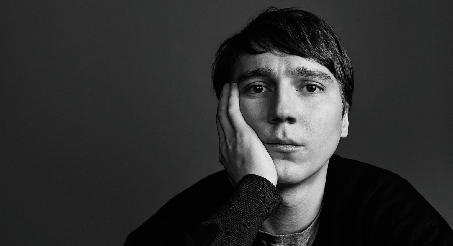 Monochrome portrait of a pensive adult man with dark hair and a grey shirt, resting his face on his hand against a dark backdrop.