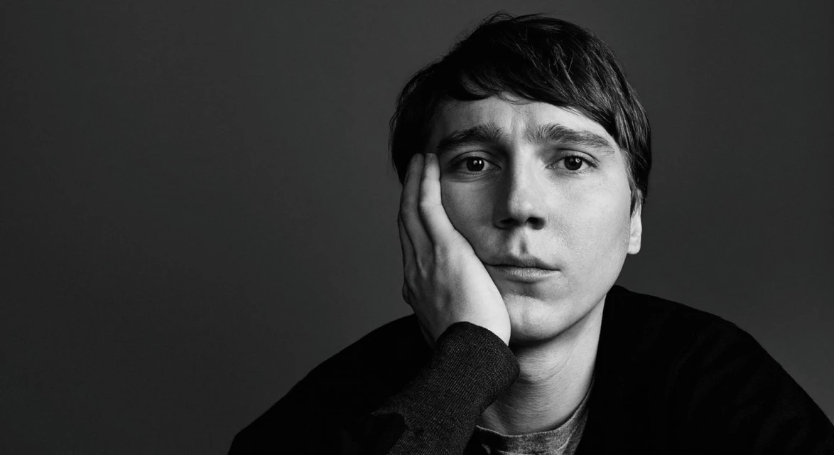 Monochrome portrait of a pensive adult man with dark hair and a grey shirt, resting his face on his hand against a dark backdrop.