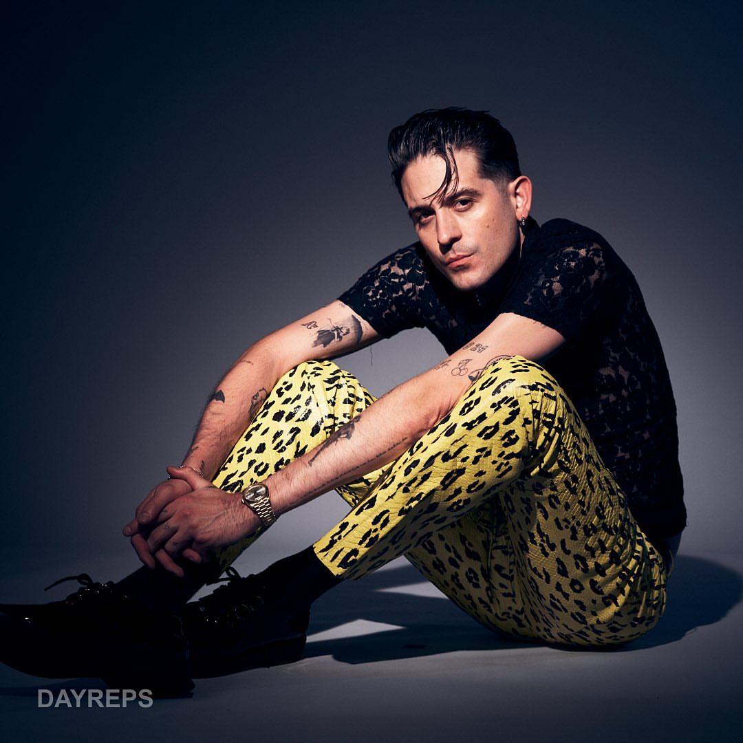 G-Eazy sitting on a grey seamless studio floor wearing yellow leopard print trousers and a black shirt