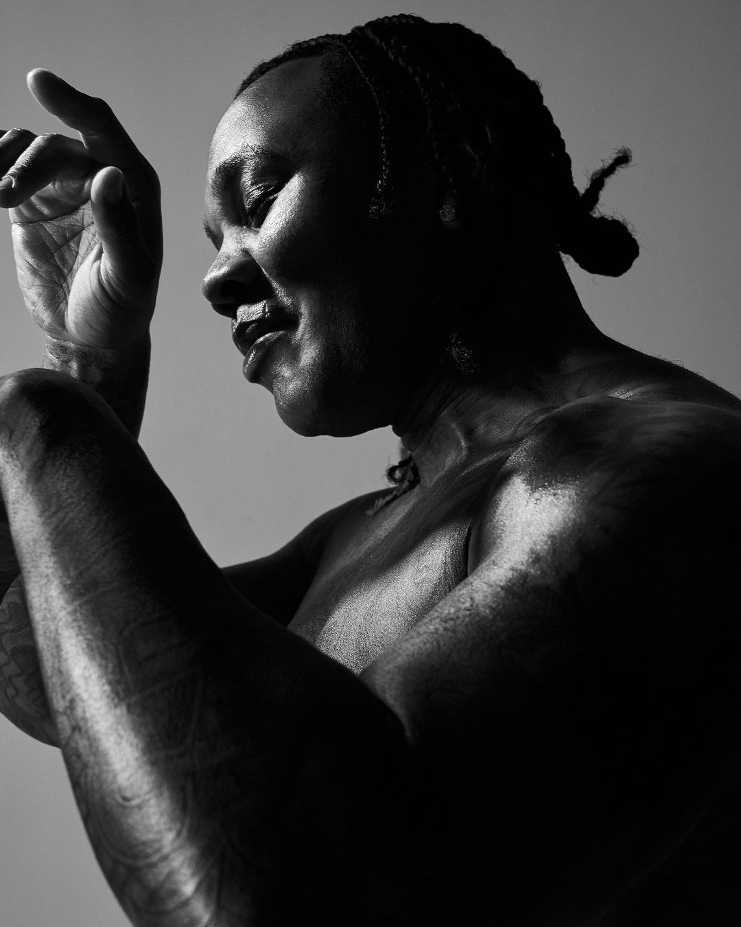 Monochrome portrait of a smiling adult with braids, facing right, arms raised, showing tattoos. The photo is shot at eye level.