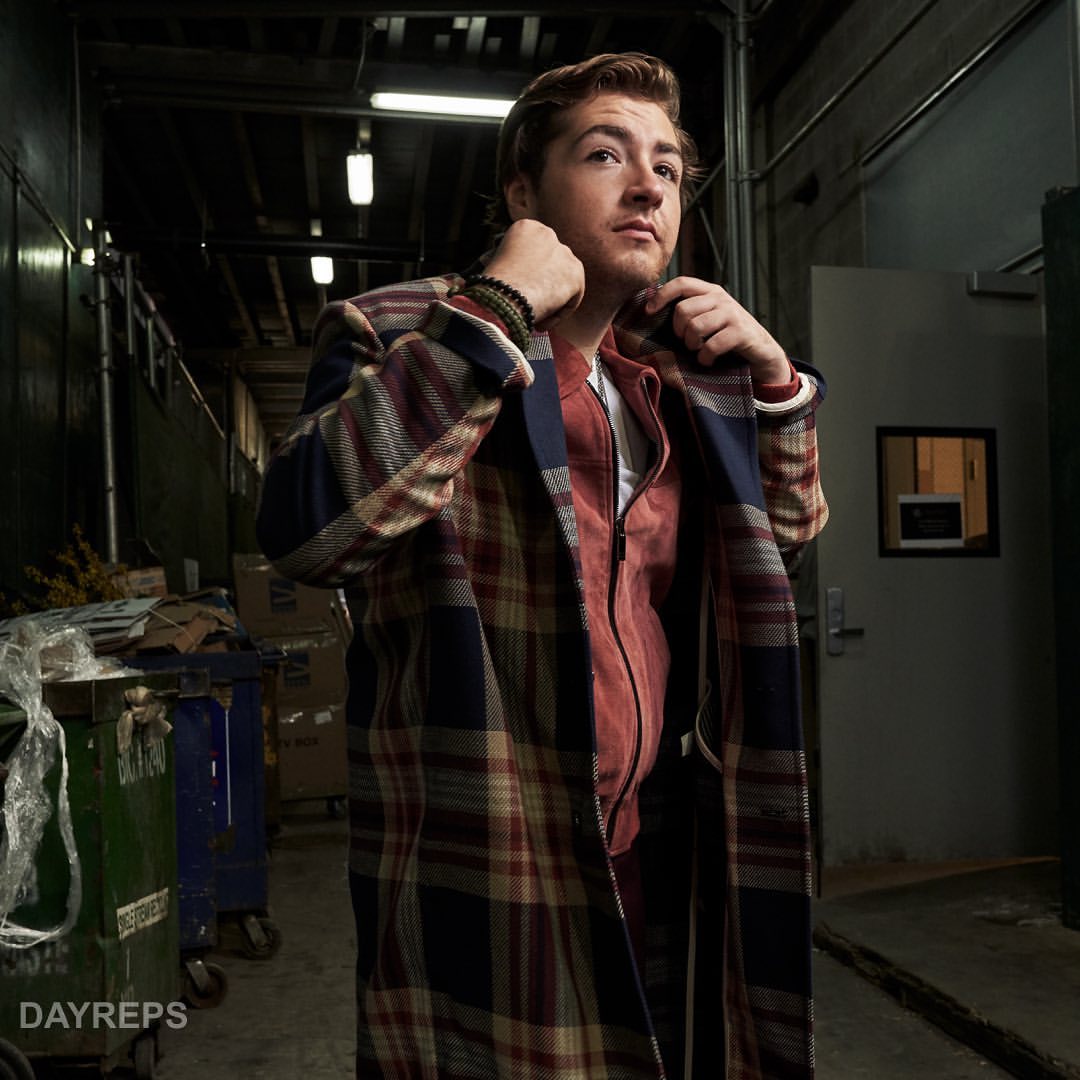 Adult man in a plaid coat stands in a long hallway with trash cans in the background, lit by overhead lights in an indoor environment.