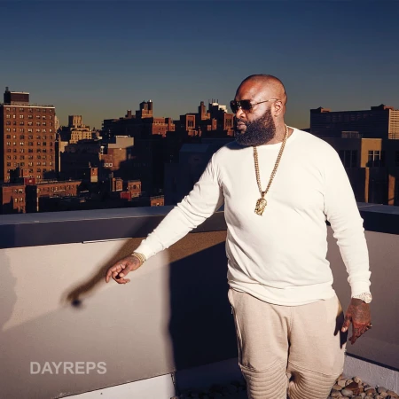 Rick Ross poses on a rooftop with city buildings in the background. He is wearing sunglasses, a gold chain, and a cream-colored outfit.
