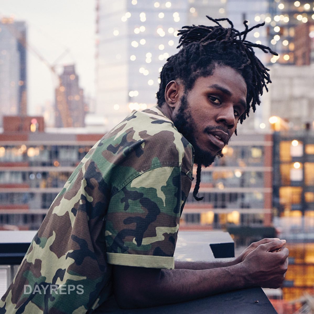 Portrait of a Black male wearing a camouflage shirt, leaning against a rail with city buildings in soft focus as background.