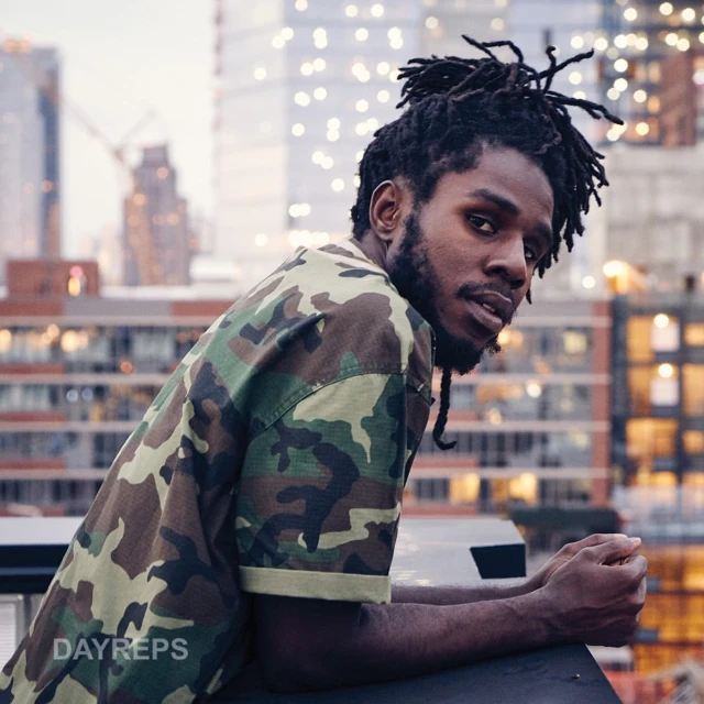 Portrait of a Black male wearing a camouflage shirt, leaning against a rail with city buildings in soft focus as background.