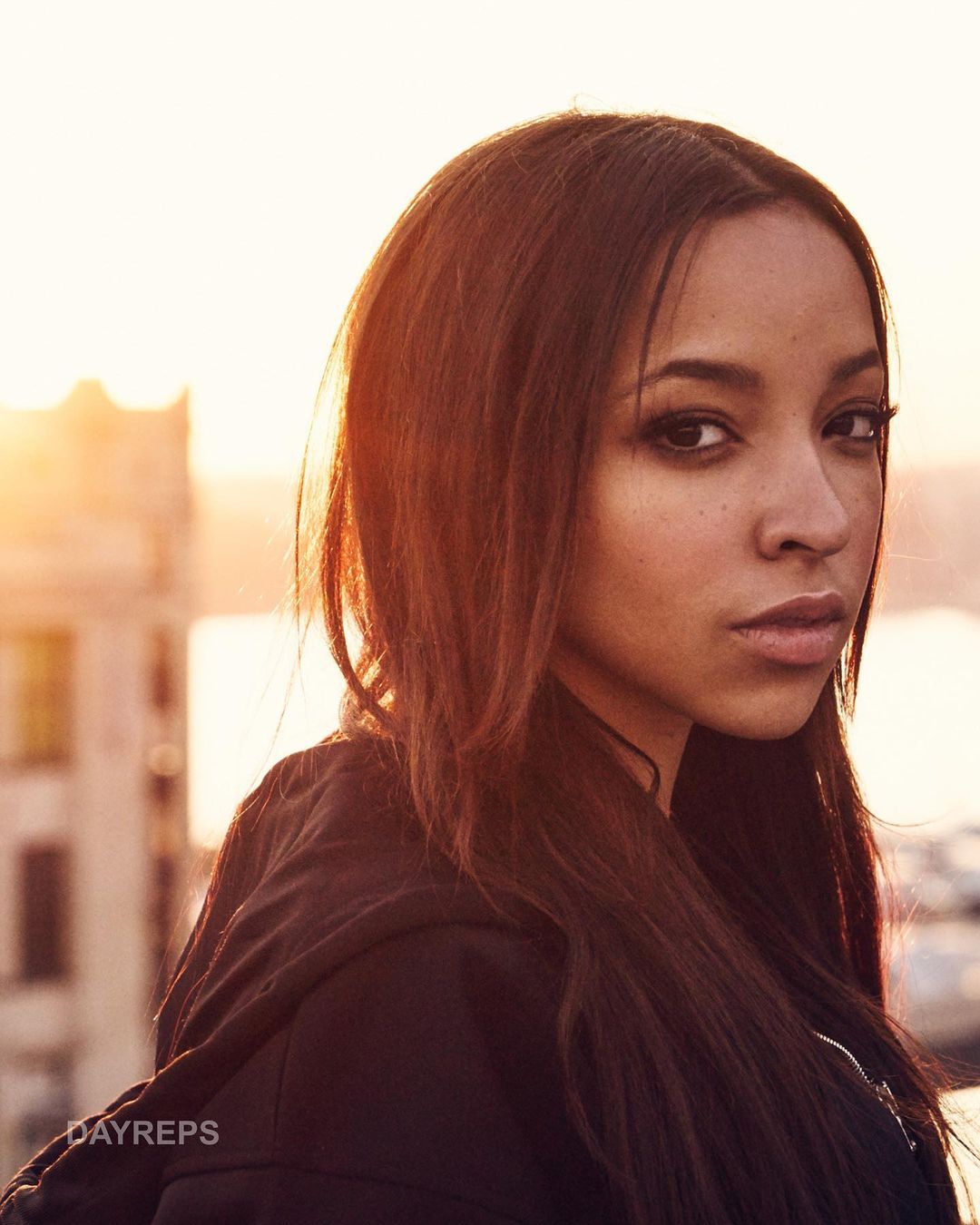 Portrait of a woman with long brown hair wearing a black jacket, shot at eye-level with a warm sunset background and urban feel.