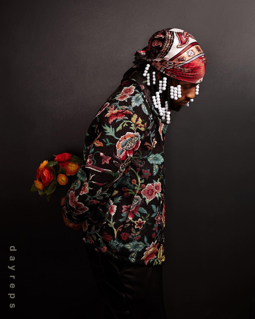Portrait of a black male model in a floral jacket and headscarf, holding flowers against a dark background in a photo studio.
