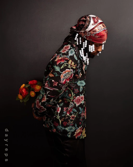 Portrait of a black male model in a floral jacket and headscarf, holding flowers against a dark background in a photo studio.