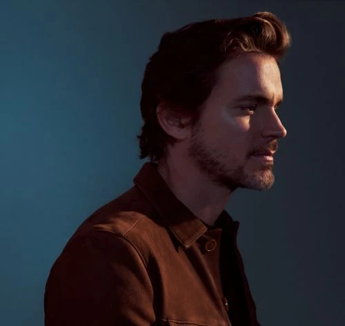 Profile of an adult male with dark hair and a brown shirt against a teal backdrop. Portrait-style, shoulder-up shot with a shallow depth of field.