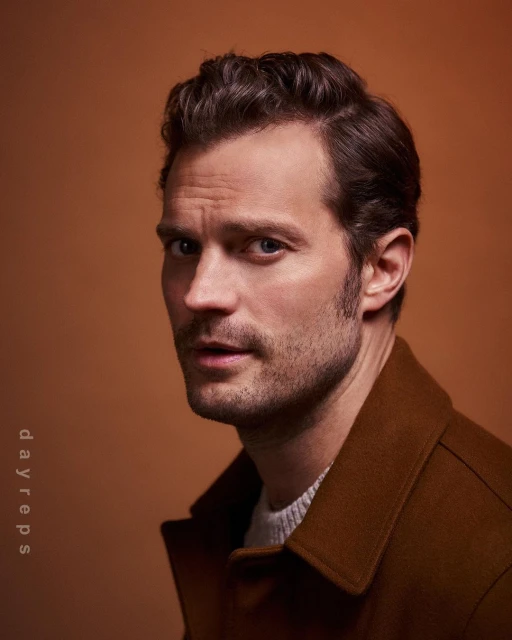 Close-up portrait of an adult male with dark hair, wearing a brown coat and a light-colored sweater, against a warm brown backdrop.