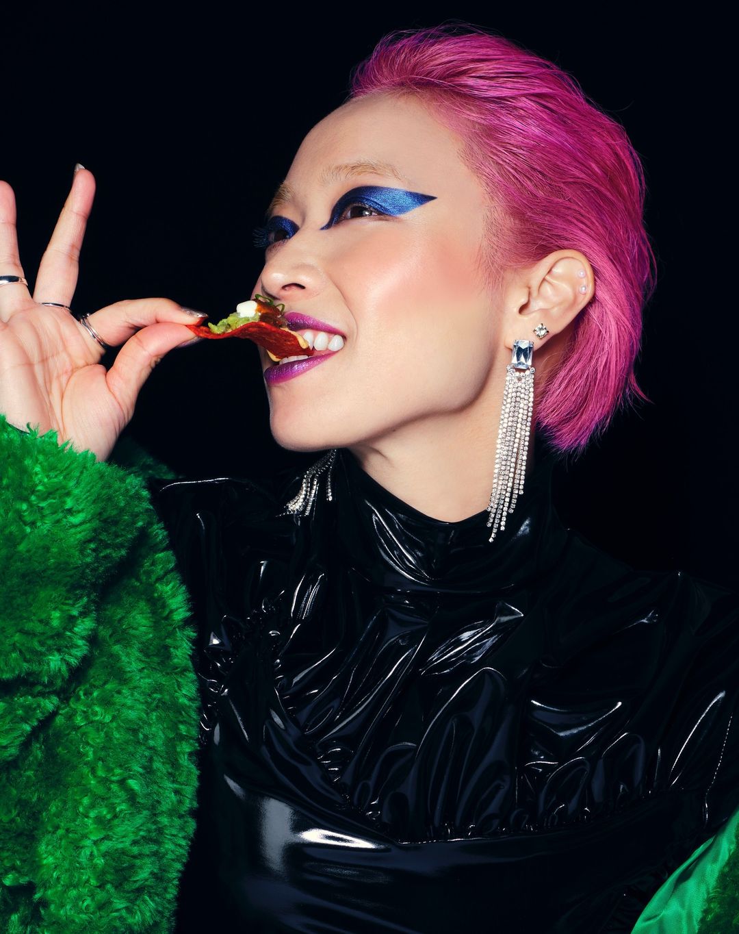 Asian woman with pink hair and blue eyeshadow, eating a pizza slice, wearing a black latex top and green fur coat against a black background.