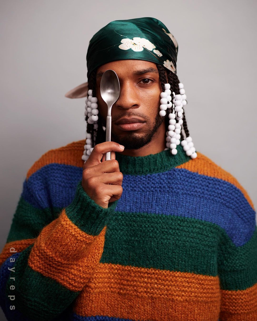 Headshot of a Black male model posing indoors with a spoon covering one eye, wearing a colorful sweater and headscarf with braids and beads.