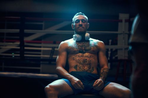 Shirtless male MMA fighter, Conor McGregor, sitting in boxing ring wearing headphones and tattoos; dark, moody atmosphere.