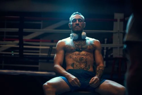 Shirtless male MMA fighter, Conor McGregor, sitting in boxing ring wearing headphones and tattoos; dark, moody atmosphere.