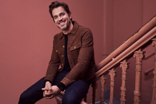 Smiling adult male with dark hair and casual brown jacket sitting on a staircase with carved wooden railings against a plain background.