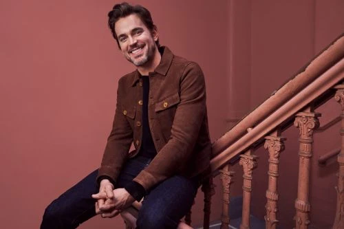 Smiling adult male with dark hair and casual brown jacket sitting on a staircase with carved wooden railings against a plain background.