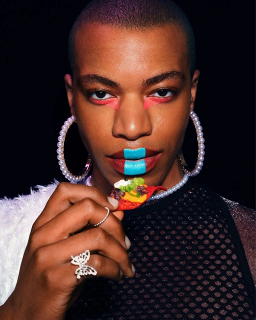 Portrait of androgynous adult person with creative makeup, pearls, fishnet top holding a filled tortilla chip in a studio with dark background.