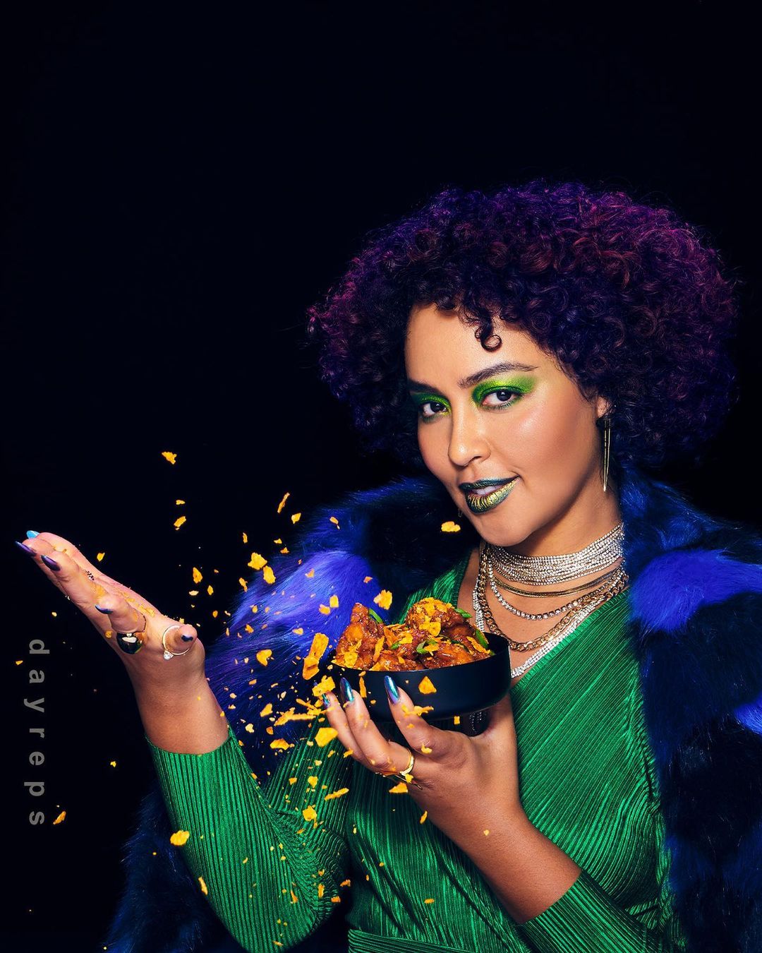 Woman in green top and faux fur holding bowl of chips, set against a dark background, showcasing beauty and food elements.
