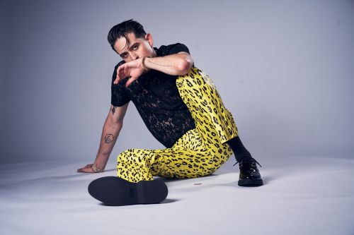 A male fashion model poses in a black lace shirt and yellow leopard-print pants, seated in front of a grey background, with a contemplative expression.