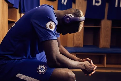 An athlete wearing headphones in a locker room looks down with his hands clasped together, dressed in blue team sportswear.