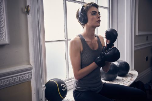 Woman in fitness wear and headphones sits on a windowsill next to medicine balls, wearing boxing gloves and looking off into the distance.
