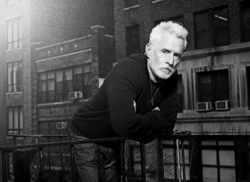 Black and white portrait of an adult man with white hair leaning on an iron railing, with brick buildings in the blurred background.