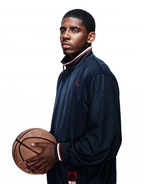 Portrait of a young adult Black man holding a basketball against a white background, wearing a blue and white track jacket.