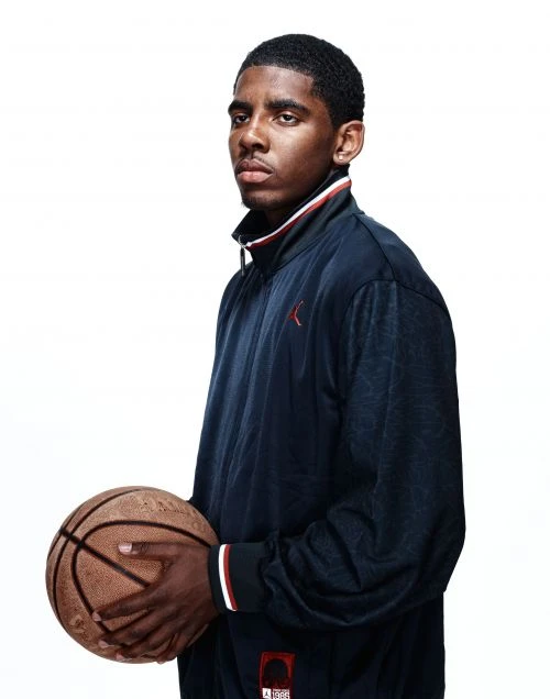 Portrait of a young adult Black man holding a basketball against a white background, wearing a blue and white track jacket.