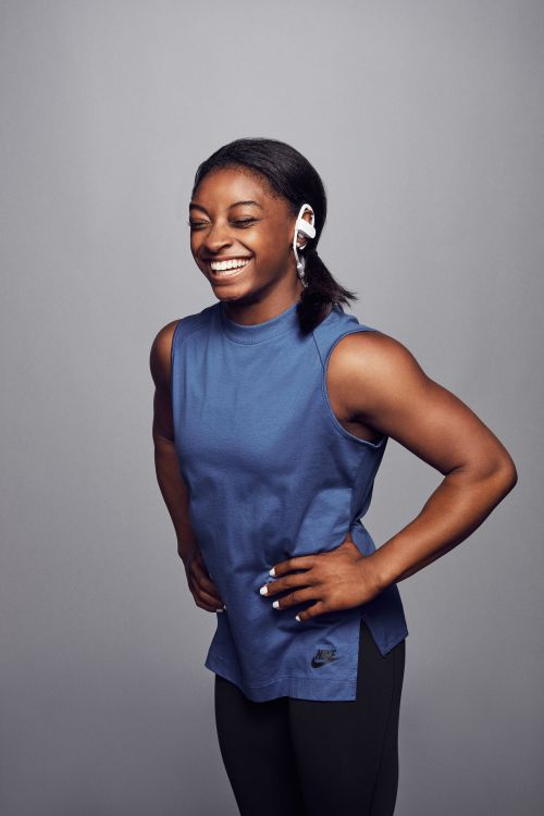 Simone Biles laughing wearing a blue sleeveless top and white headphones against a grey seamless background
