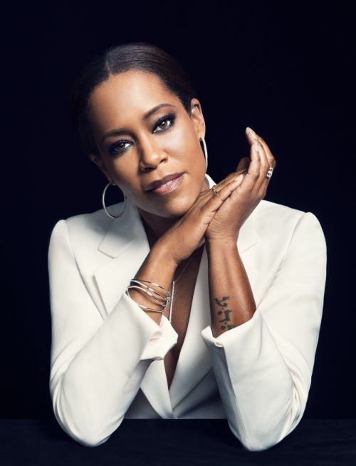 Headshot portrait of Regina King in a white blazer, gazing at the camera with a serious expression, set against a dark background.