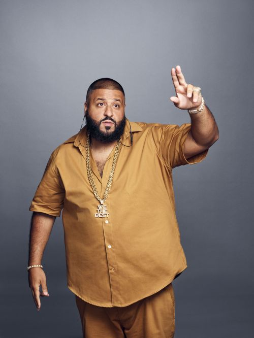 Studio portrait of DJ Khaled wearing gold jewelry, dressed in a matching gold button-down shirt and pants, standing against a gray background.