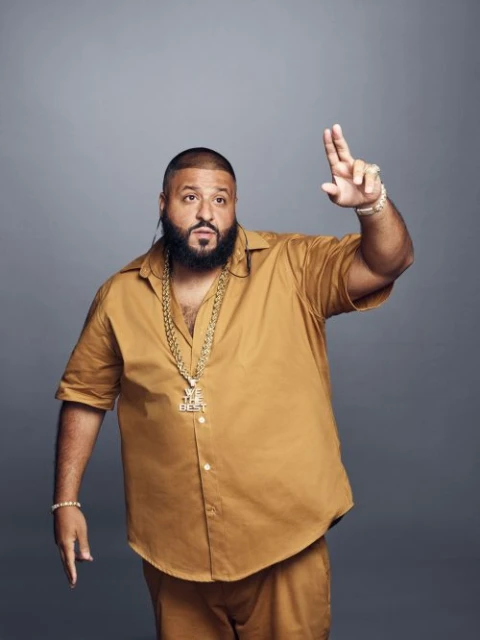 Studio portrait of DJ Khaled wearing gold jewelry, dressed in a matching gold button-down shirt and pants, standing against a gray background.