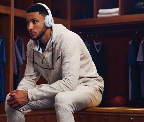 Male athlete wearing headphones and a grey tracksuit sitting in a locker room with lockers and a basketball in the background.