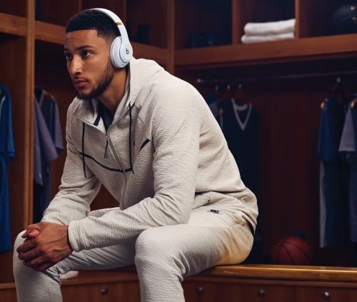 Male athlete wearing headphones and a grey tracksuit sitting in a locker room with lockers and a basketball in the background.