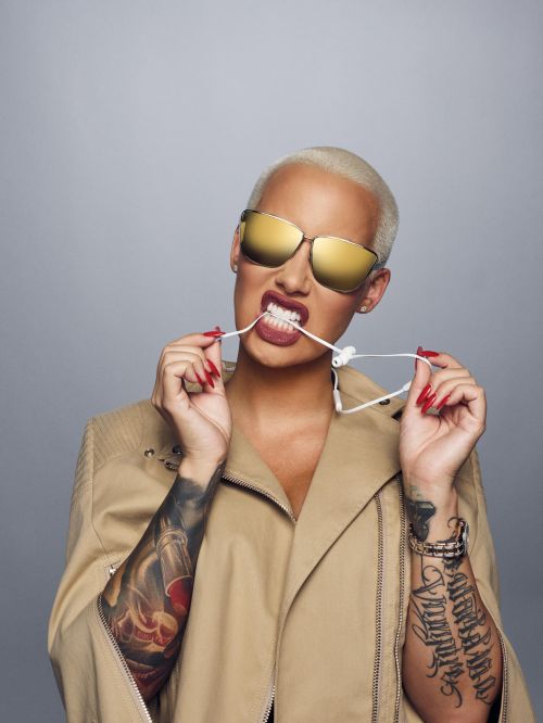 Portrait of Amber Rose, wearing gold sunglasses and a tan jacket, biting the cord of white earbuds against a light gray backdrop.