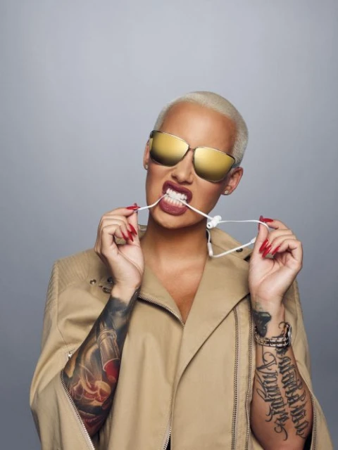 Portrait of Amber Rose, wearing gold sunglasses and a tan jacket, biting the cord of white earbuds against a light gray backdrop.
