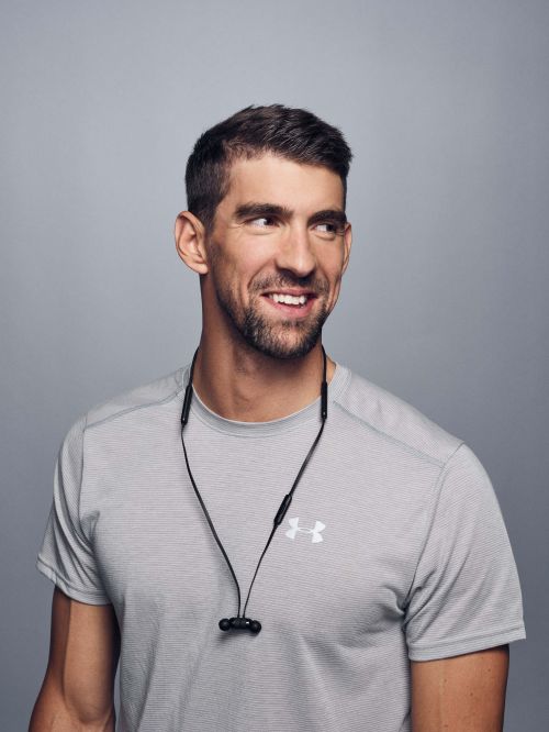 Portrait of Michael Phelps smiling and looking away from the camera with wired earbuds around his neck in a photo studio setting.