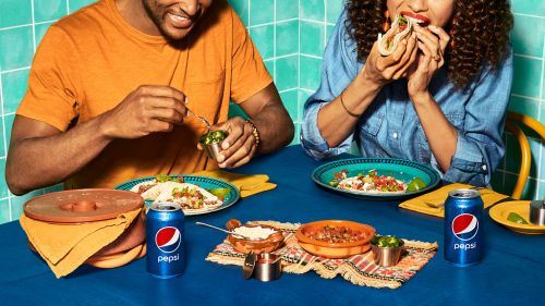 Two people enjoy tacos and Pepsi at a table, featuring vibrant food, a colorful tablecloth, and refreshing drinks in an indoor setting.